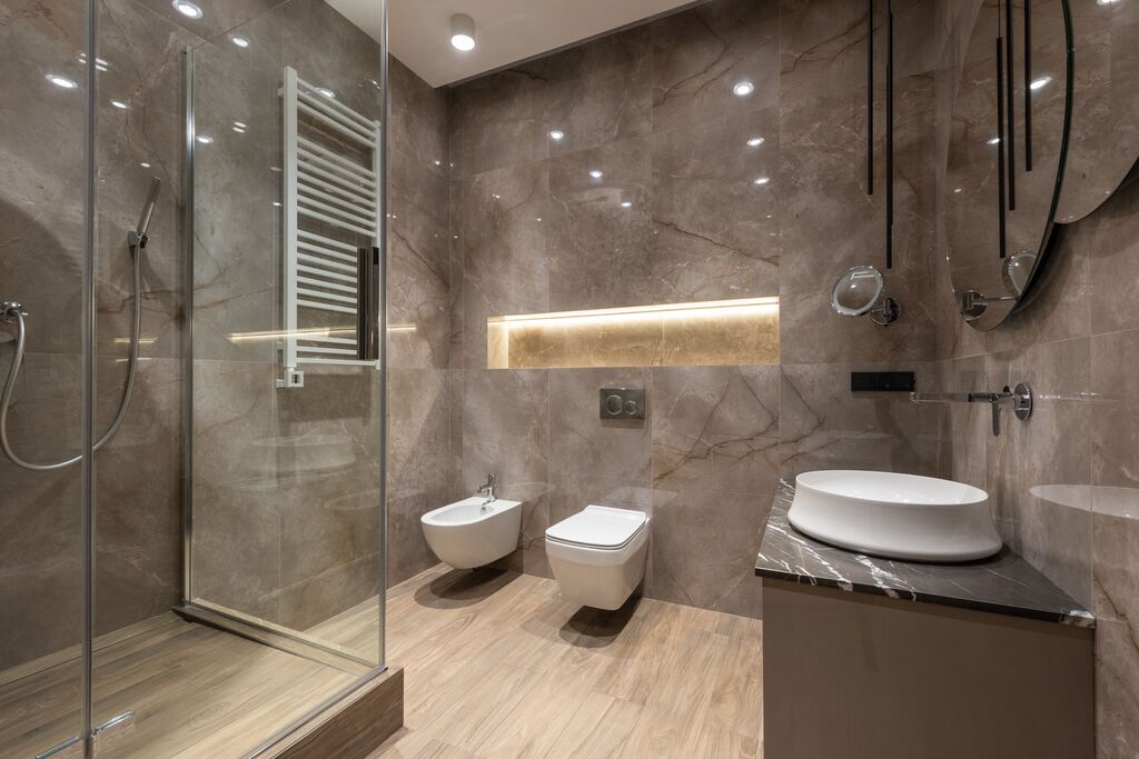 Brown tiled bathroom with wooden floors and two white toilets, a clear shower, and a white basin.